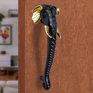 May include: Black elephant-shaped door handle with gold accents. The handle features a detailed elephant head with gold ears and tusks. The handle's body is black and has a textured, curved design. The handle is mounted on a brown wooden door.