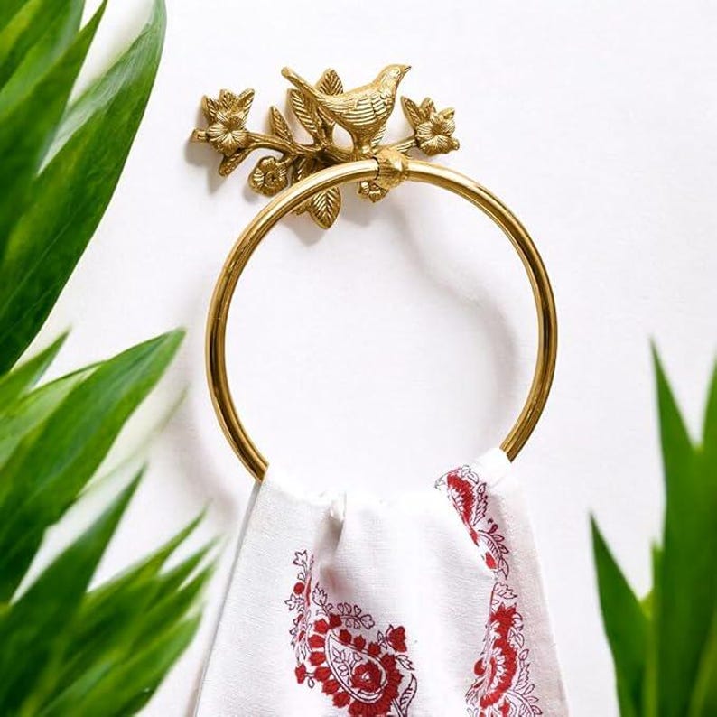 Puede incluir: Un toallero dorado con un dise&ntilde;o de p&aacute;jaro y flores. El anillo sostiene una toalla blanca con un estampado floral rojo. El toallero est&aacute; montado en una pared blanca, con hojas verdes en el fondo.