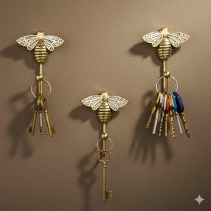 May include: Three brass bee-shaped key holders on a brown wall. Each has a detailed bee design with white wing accents. Various keys hang from the holders, adding functionality and decorative appeal. The holders are a stylish home decor item.