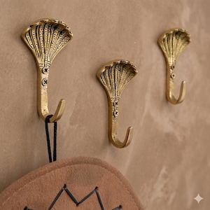 Peut inclure: Trois crochets muraux dorés en forme de têtes de cobra sont fixés sur un mur beige. Les crochets présentent un motif détaillé et texturé et sont vissés au mur. Un chapeau marron avec des coutures noires est suspendu à l'un des crochets.