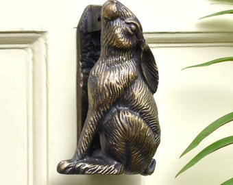 Könnte beinhalten: Türklopfer in Bronzeoptik in Form eines sitzenden Hasen. Der Klopfer ist auf einer dunklen rechteckigen Platte montiert und an einer cremefarbenen Tür befestigt. Der Hase ist mit strukturierter Fell versehen und hat den Kopf nach oben geneigt.