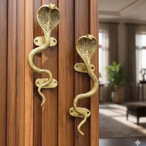 Peut inclure: Deux poignées de porte en laiton en forme de cobra, montées sur une porte en bois. Les poignées présentent des motifs d'écailles détaillés et un corps incurvé. Les têtes de cobra sont en haut. Les poignées sont de couleur dorée.