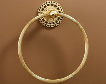 May include: A gold-colored towel ring with a decorative circular backplate. The ring is round with a ridged texture, and the backplate features a detailed, openwork design. The fixture is mounted on a brown surface.