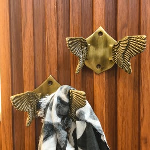 Może przedstawiać: Dwa złote metalowe haczyki ścienne z wzorem skrzydeł. Na jednym haczyku wisi szalik.