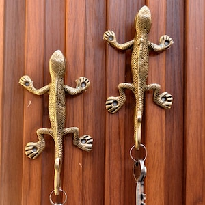 Könnte beinhalten: Zwei goldfarbene Schlüsselhaken in Form einer Eidechse, die an einer Holzwand montiert sind. Die Haken haben ein detailliertes Design und eignen sich perfekt zum Aufhängen von Schlüsseln oder anderen kleinen Gegenständen.