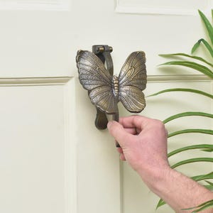 Puede incluir: Un aldaba de puerta con forma de mariposa de bronce. La aldaba est&aacute; montada en una puerta de color crema. La mariposa tiene patrones de alas detallados y un cuerpo central. Una mano sostiene la aldaba.