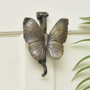 Puede incluir: Un llamador de puerta con forma de mariposa en color bronce. Las alas est&aacute;n detalladas con patrones de venas. Est&aacute; montado en una puerta blanca. Un elemento decorativo para el hogar.