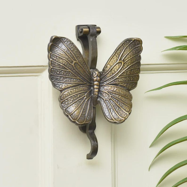 Puede incluir: Un llamador de puerta con forma de mariposa en color bronce. Las alas est&aacute;n detalladas con patrones de venas. Est&aacute; montado en una puerta blanca. Un elemento decorativo para el hogar.
