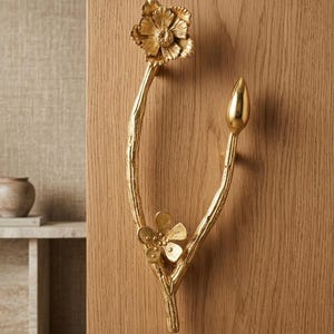 Könnte beinhalten: Ein goldfarbener Türgriff mit floralem Design, bestehend aus einer Blüte und einer Knospe. Der Griff ist an einer Holztür befestigt und zeigt seine filigranen Details und seine elegante Ästhetik. Das Design umfasst eine große Blüte oben und eine kleinere Blüte unten.