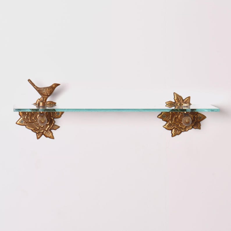 Puede incluir: Una estanter&iacute;a decorativa de cristal con detalles florales y de p&aacute;jaros de color lat&oacute;n. La estanter&iacute;a est&aacute; sostenida por dos soportes ornamentados, uno con un p&aacute;jaro posado sobre una base floral y otro con un dise&ntilde;o floral. La estanter&iacute;a mide unos 61 cm de largo.