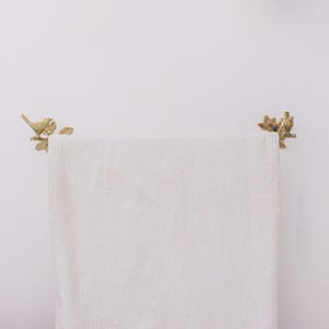 Puede incluir: Una toalla blanca con relieve cuelga de una barra de toalla decorativa de lat&oacute;n. La barra de la toalla presenta dise&ntilde;os de p&aacute;jaros y hojas en cada extremo. El fondo es una pared blanca lisa.