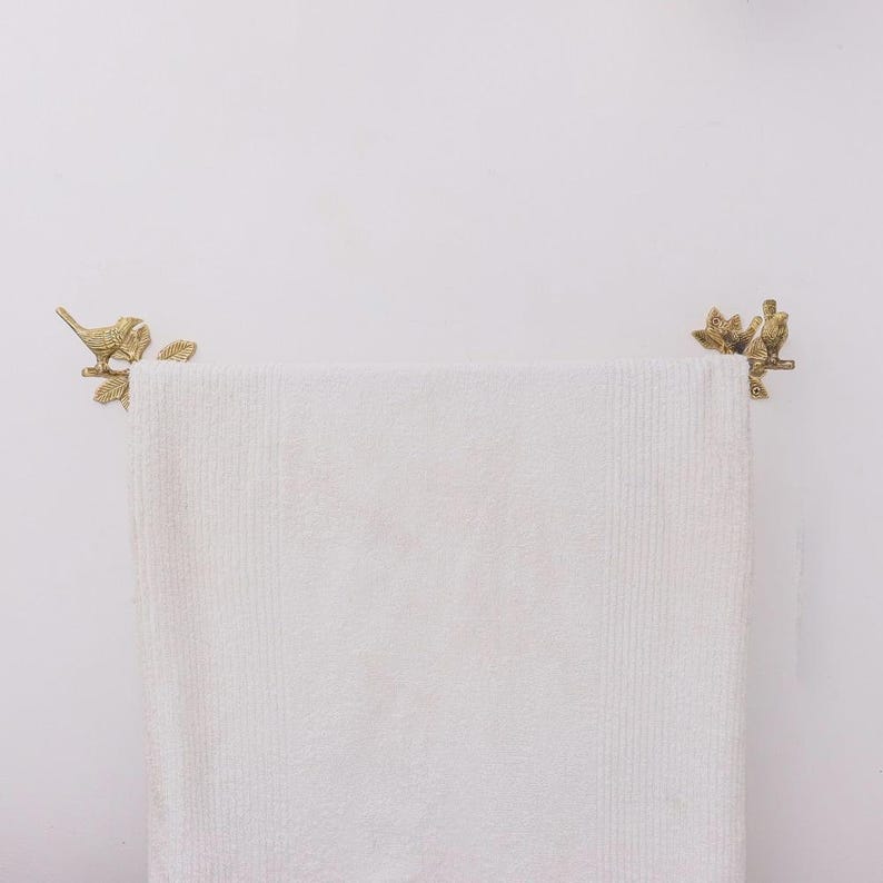 Puede incluir: Una toalla blanca con relieve cuelga de una barra de toalla decorativa de lat&oacute;n. La barra de la toalla presenta dise&ntilde;os de p&aacute;jaros y hojas en cada extremo. El fondo es una pared blanca lisa.