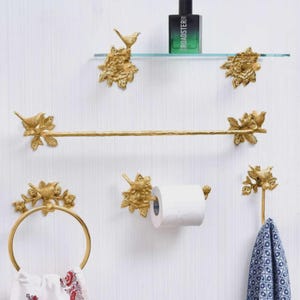 Puede incluir: Un conjunto de accesorios de ba&ntilde;o dorados, que incluyen una barra para toallas, un aro para toallas, un portarrollos de papel higi&eacute;nico, soportes para estantes y un gancho. Cada pieza presenta un dise&ntilde;o de p&aacute;jaros y flores. Una toalla azul y blanca cuelga del gancho.