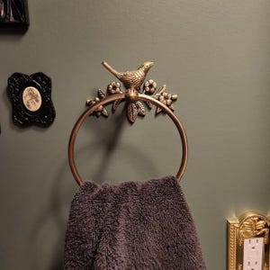 Puede incluir: Un toallero de color bronce con un dise&ntilde;o decorativo de p&aacute;jaro y flores. Sostiene una toalla gris oscuro. La pared est&aacute; pintada de color verde oscuro. Tambi&eacute;n se ven marcos decorativos y una cubierta dorada para enchufes.