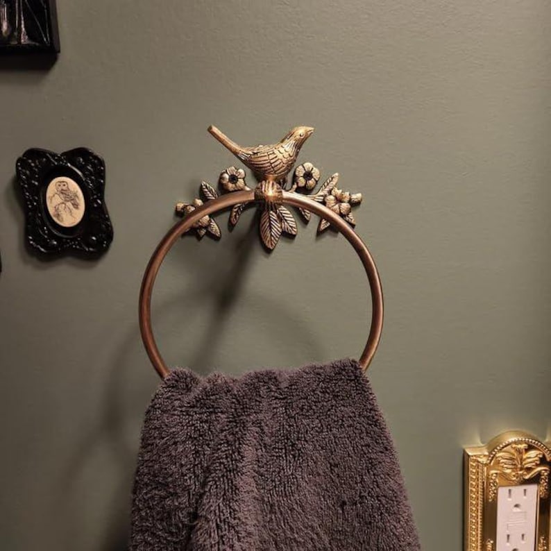 Puede incluir: Un toallero de color bronce con un dise&ntilde;o decorativo de p&aacute;jaro y flores. Sostiene una toalla gris oscuro. La pared est&aacute; pintada de color verde oscuro. Tambi&eacute;n se ven marcos decorativos y una cubierta dorada para enchufes.
