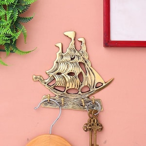 Puede incluir: Un llavero de pared de metal dorado con forma de velero. El llavero tiene varios ganchos para llaves y un diseño decorativo. El velero está montado en una pared rosa, con una percha de madera y llaves colgando de los ganchos.