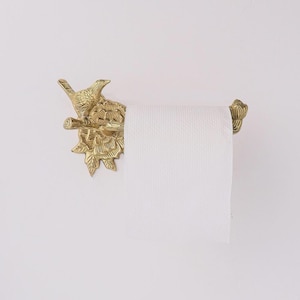 Puede incluir: Un portarrollos de papel higi&eacute;nico de color lat&oacute;n con un p&aacute;jaro y hojas. El soporte est&aacute; montado en una pared blanca y contiene un rollo de papel higi&eacute;nico blanco. El dise&ntilde;o ornamentado a&ntilde;ade un toque decorativo a un ba&ntilde;o.