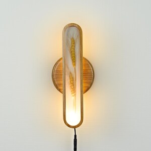 Pode incluir: Uma luminária de parede com uma moldura de madeira e um inserto de vidro transparente. O inserto de vidro apresenta um caule de trigo seco. A luz está acesa e emite um brilho quente.