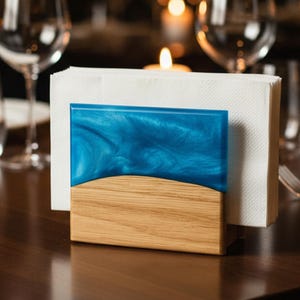 May include: A wooden and blue resin napkin holder on a dark wood table. The holder has a rectangular shape with a blue, swirled resin top and a wooden base. White napkins are in the holder. Wine glasses and silverware are in the background.