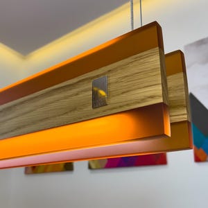 May include: A linear pendant light fixture featuring a horizontal wooden beam with a prominent grain pattern, accented by translucent orange resin panels. A small, rectangular metallic plate is embedded on the side of the wood section.