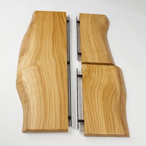May include: Four wooden serving boards with a natural edge and a light brown finish. The boards are connected by black metal supports. The wood grain is visible, and the boards are arranged on a white surface.