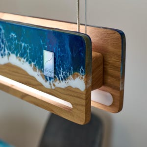 May include: A wooden pendant light fixture with a beach scene design. The fixture features a rectangular shape with a light strip and a resin panel depicting ocean waves in shades of blue and white. The fixture has a wooden frame and a logo that says "Hedonism Wood".