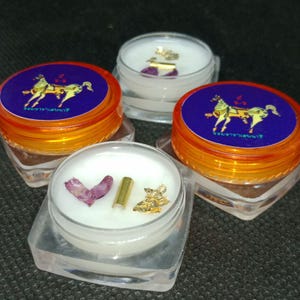 May include: Four small, clear, square containers with white contents, gold flakes, and dried rose petals. The containers have orange lids with a blue label featuring a gold horse illustration. The items are arranged on a dark surface.