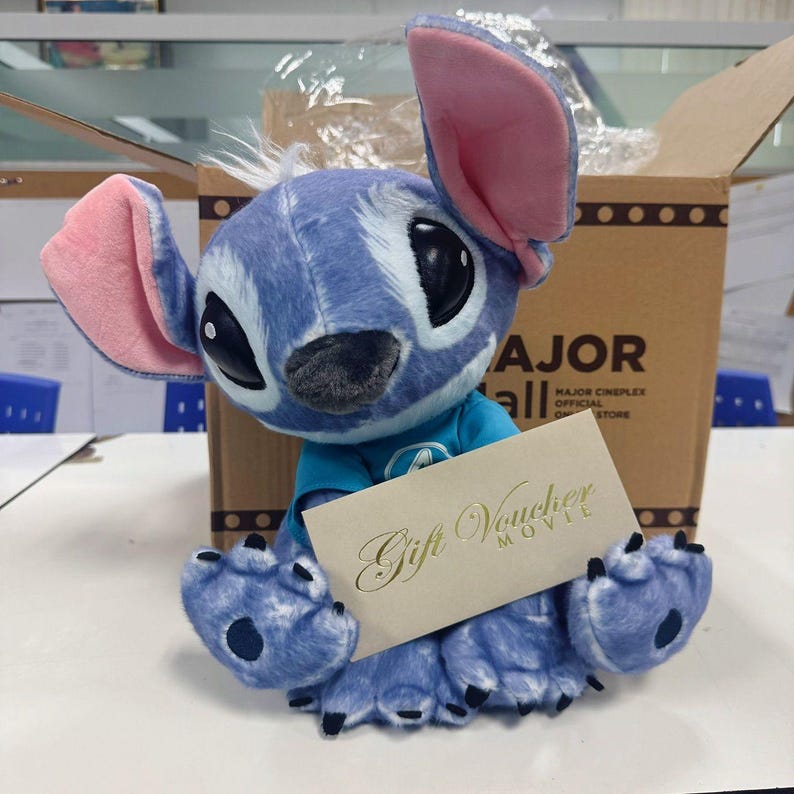 May include: A blue plush Stitch toy with large black eyes and pink ears, holding a gift voucher. The toy is wearing a blue top and is in front of a cardboard box. The gift voucher reads 'Gift Voucher MOVIE'.