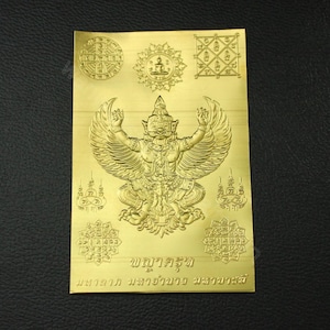 May include: A gold-colored metal plaque with a raised image of a mythical bird-like creature with outstretched wings. The creature is surrounded by various symbols and text in Thai. The text at the bottom reads "ชฎาครุฑ มหาอำนาจ มหาโชคลาภ".
