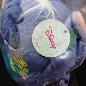 May include: A blue plush toy of a Disney character, possibly Stitch, in a clear plastic bag. The toy has white and yellow details. A Disney-branded tag is visible.