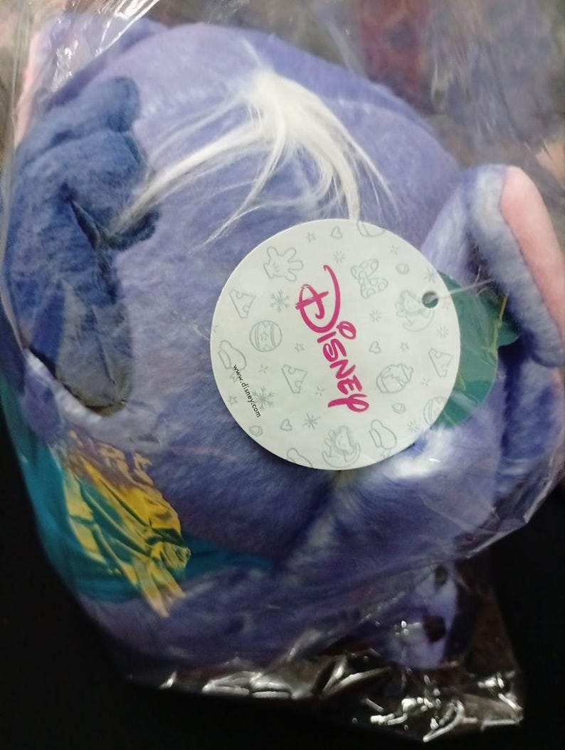 May include: A blue plush toy of a Disney character, possibly Stitch, in a clear plastic bag. The toy has white and yellow details. A Disney-branded tag is visible.