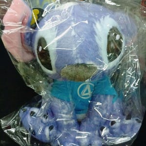 May include: A soft toy of a blue alien-like creature with large eyes, pink ears, and a blue shirt with a white circle and a blue number 4. The toy is in a clear plastic bag.