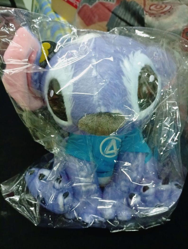May include: A soft toy of a blue alien-like creature with large eyes, pink ears, and a blue shirt with a white circle and a blue number 4. The toy is in a clear plastic bag.
