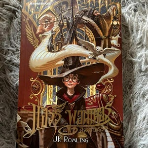 May include: A burgundy-covered hardcover book with an illustrated cover. The artwork shows a young person with glasses and a hat, set against a backdrop of fantastical imagery. The title is in a decorative font, with the author's name, J.K. Rowling, beneath.