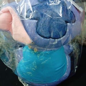 May include: A soft toy of a blue alien character with large ears and a turquoise belly, packaged in a clear plastic bag. The character has a dark blue patch on its head and pink inner ears. The toy is soft and fluffy.
