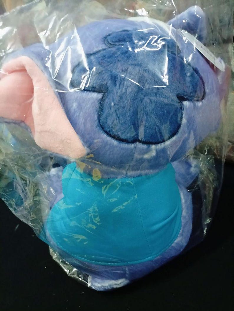 May include: A soft toy of a blue alien character with large ears and a turquoise belly, packaged in a clear plastic bag. The character has a dark blue patch on its head and pink inner ears. The toy is soft and fluffy.