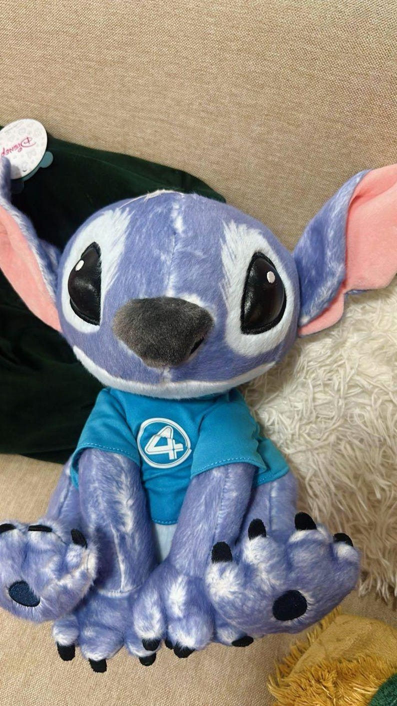 May include: A soft toy of a blue alien-like character with large ears and black eyes. It is wearing a blue top with a white number 4. The toy is light blue with darker blue spots.