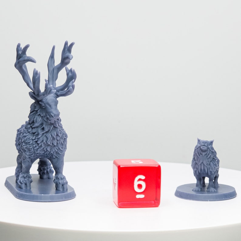 May include: Three miniature figurines: a detailed gray stag with large antlers, a small gray wolf, and a red die with the number 6 visible. The stag and wolf are on gray bases. The die is a cube with white numbers.