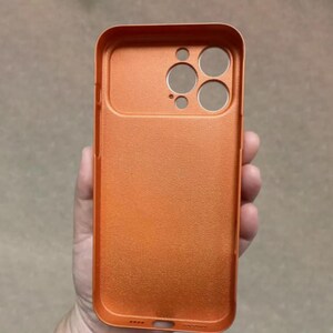 May include: Orange phone case with a textured surface. The case has cutouts for the camera and other phone features. The case is held in a hand.