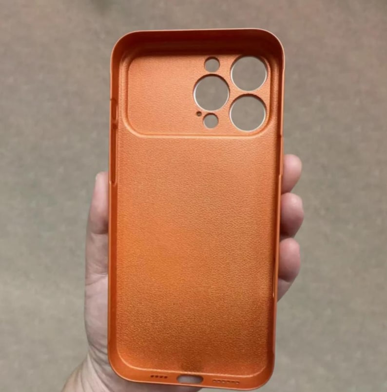 May include: Orange phone case with a textured surface. The case has cutouts for the camera and other phone features. The case is held in a hand.