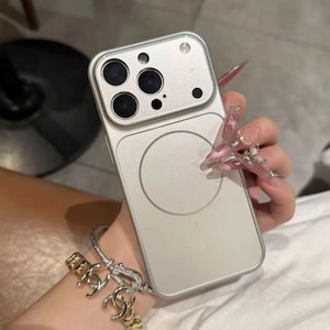 May include: A silver phone case with a magnetic circle on the back. The case has a camera cutout with multiple lenses. The phone case is held by a person wearing a bracelet.