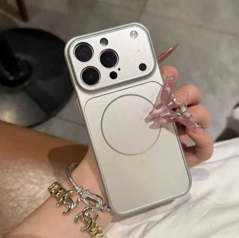 May include: A silver phone case with a magnetic circle on the back. The case has a camera cutout with multiple lenses. The phone case is held by a person wearing a bracelet.