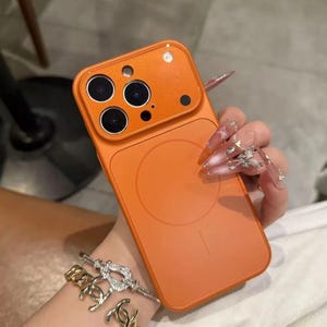 May include: Orange phone case with a magnetic circle on the back. The case has a camera cutout with three lenses and a flash. The case is held by a person with long, decorated nails. A gold and silver bracelet is also visible.