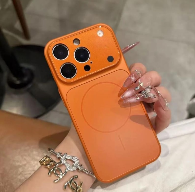 May include: Orange phone case with a magnetic circle on the back. The case has a camera cutout with three lenses and a flash. The case is held by a person with long, decorated nails. A gold and silver bracelet is also visible.