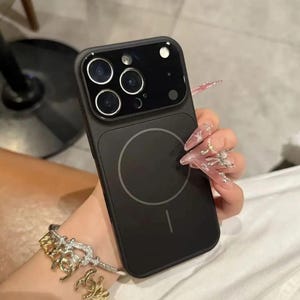 May include: Black phone case with a circular design on the back and a camera module with three lenses. The case is held in a hand with long, decorated nails. A gold and silver bracelet is visible.