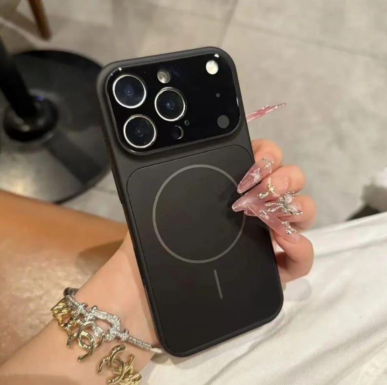May include: Black phone case with a circular design on the back and a camera module with three lenses. The case is held in a hand with long, decorated nails. A gold and silver bracelet is visible.