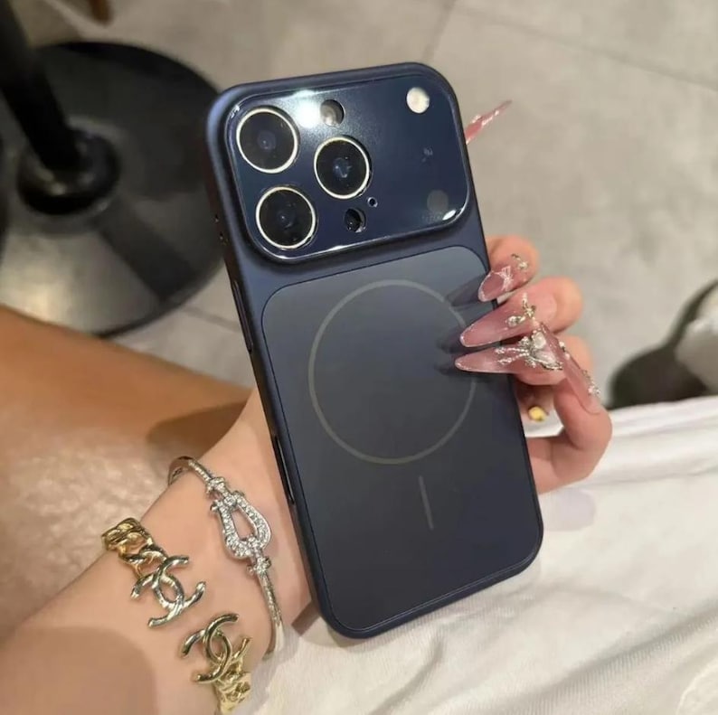 May include: A dark blue phone case with a circular design on the back, held in a hand with long, decorated nails. The case has a camera module with three lenses and a flash. A gold bracelet and a silver bracelet are also visible.