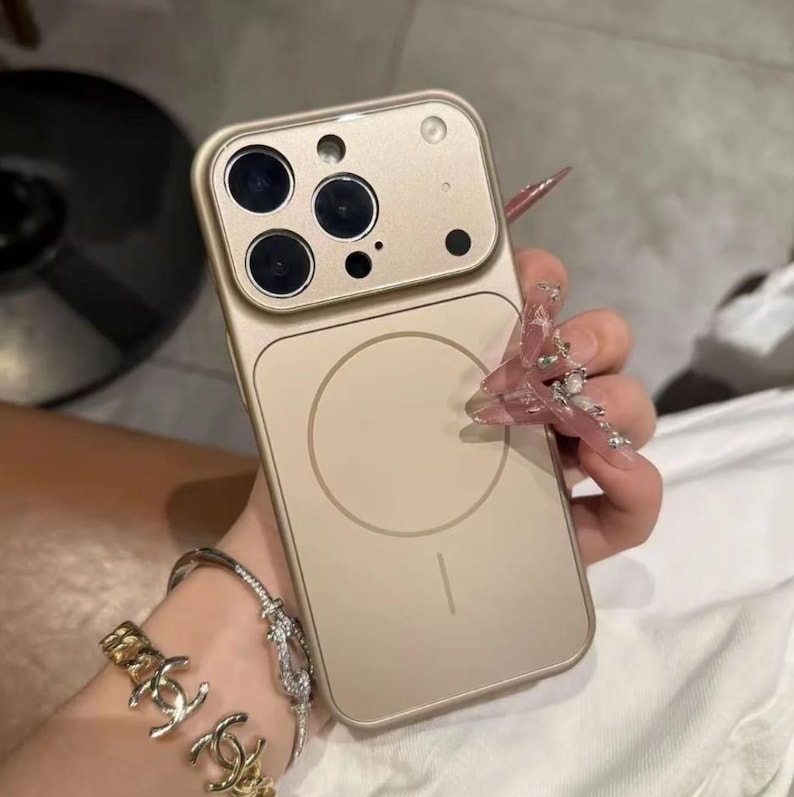 May include: A gold-colored phone case with a camera module featuring multiple lenses. The case has a circular design on the back and is held by a person wearing bracelets. The case is designed to fit a smartphone.