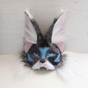 Peut inclure: Un masque de chat fait à la main avec un visage gris et noir, des accents bleus et de grandes oreilles blanches et grises duveteuses. Le masque a des yeux bleus et un nez rose. Le masque est fait de matériaux doux et est conçu pour être porté.