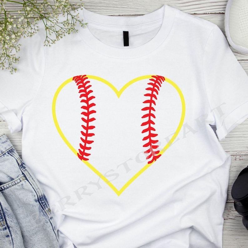 以下が含まれることがあります： 黄色のハート型の野球のグラフィックが描かれた白いTシャツ。野球は赤いステッチです。